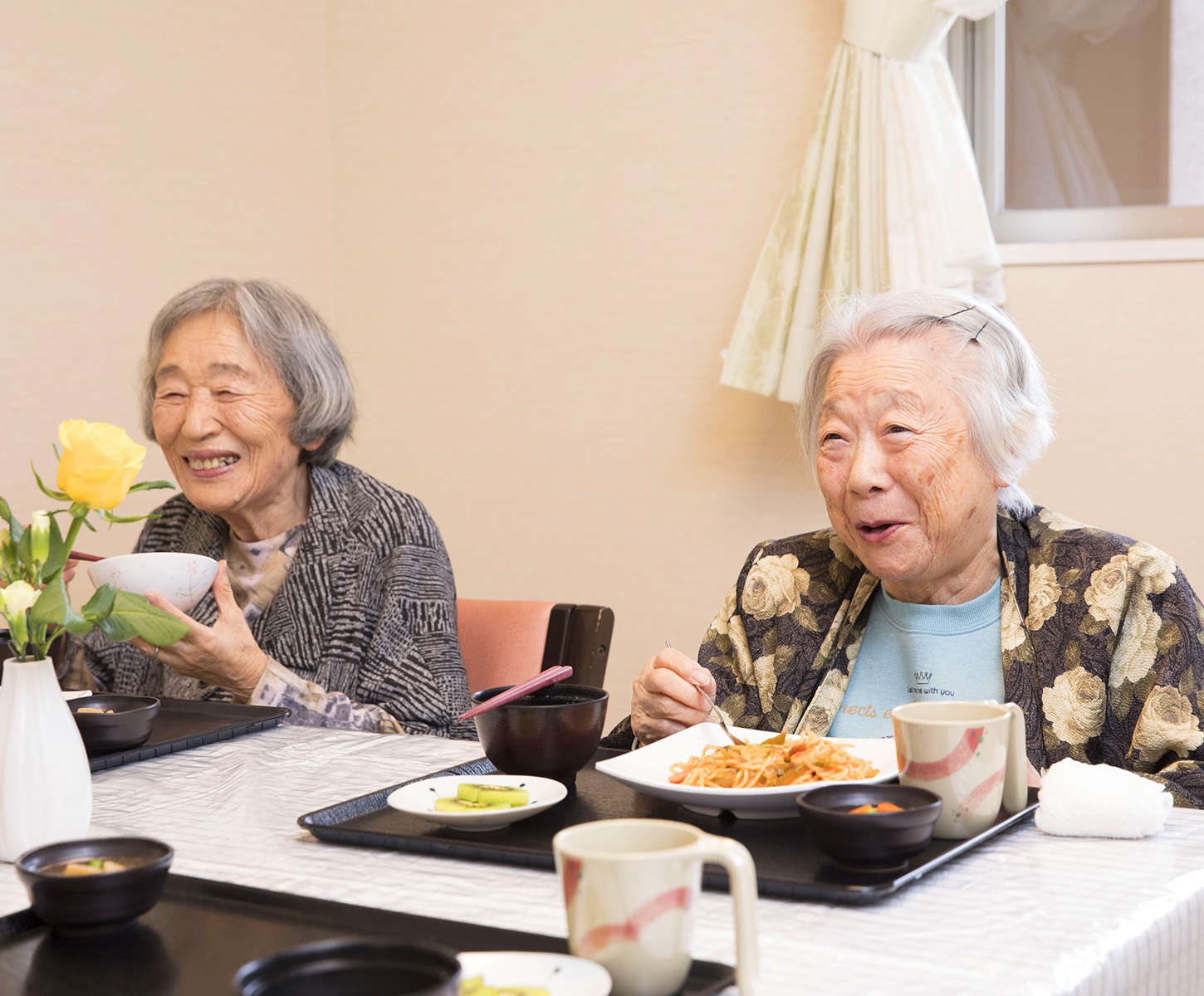 食堂で食事をする高齢入居者2名。黄色い花が飾られた温かみのある空間